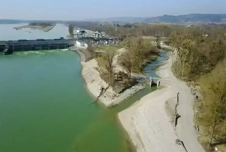 Aerial view of a power plant on a river with backed-up water, adjacent wooded area, and hiking trails in clear weather.