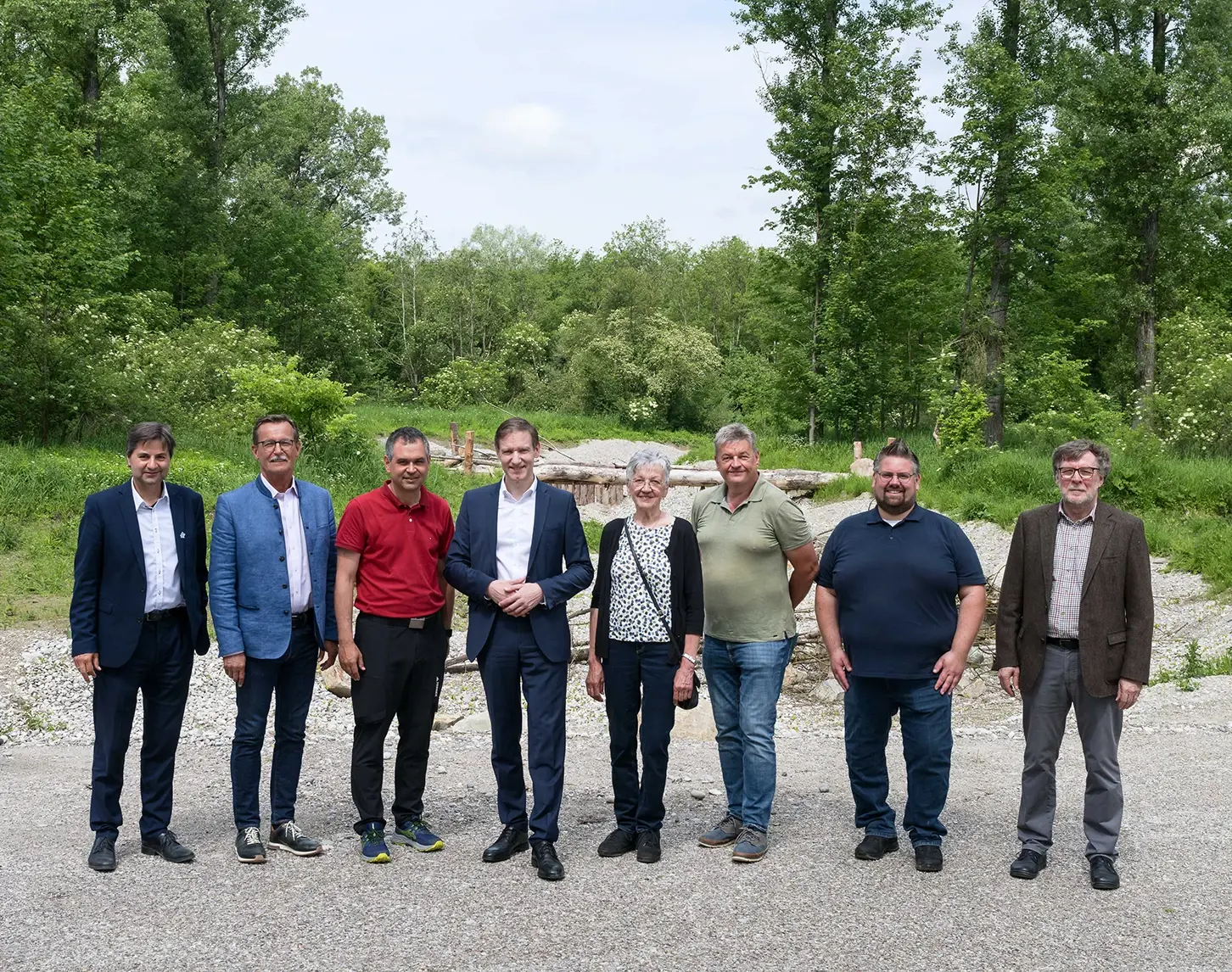 Gruppe von acht Personen in formeller und legerer Kleidung, die auf Schotterboden vor einer natürlichen Waldlandschaft stehen.