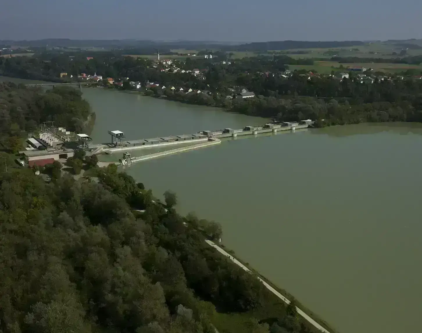 Luftaufnahme eines Laufkraftwerks, umgeben von grüner Vegetation und einer kleinen Ortschaft im Hintergrund.