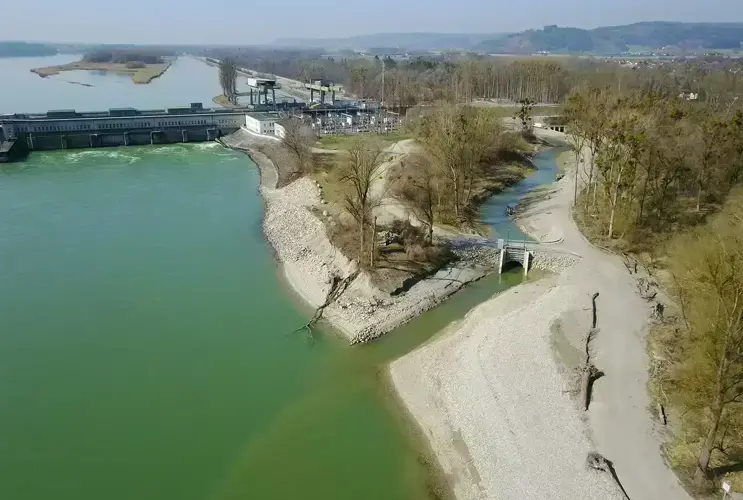 Luftaufnahme eines Kraftwerks an einem Fluss mit aufgestautem Wasser, angrenzendem Waldgebiet und Wanderwegen bei klarem Wetter.