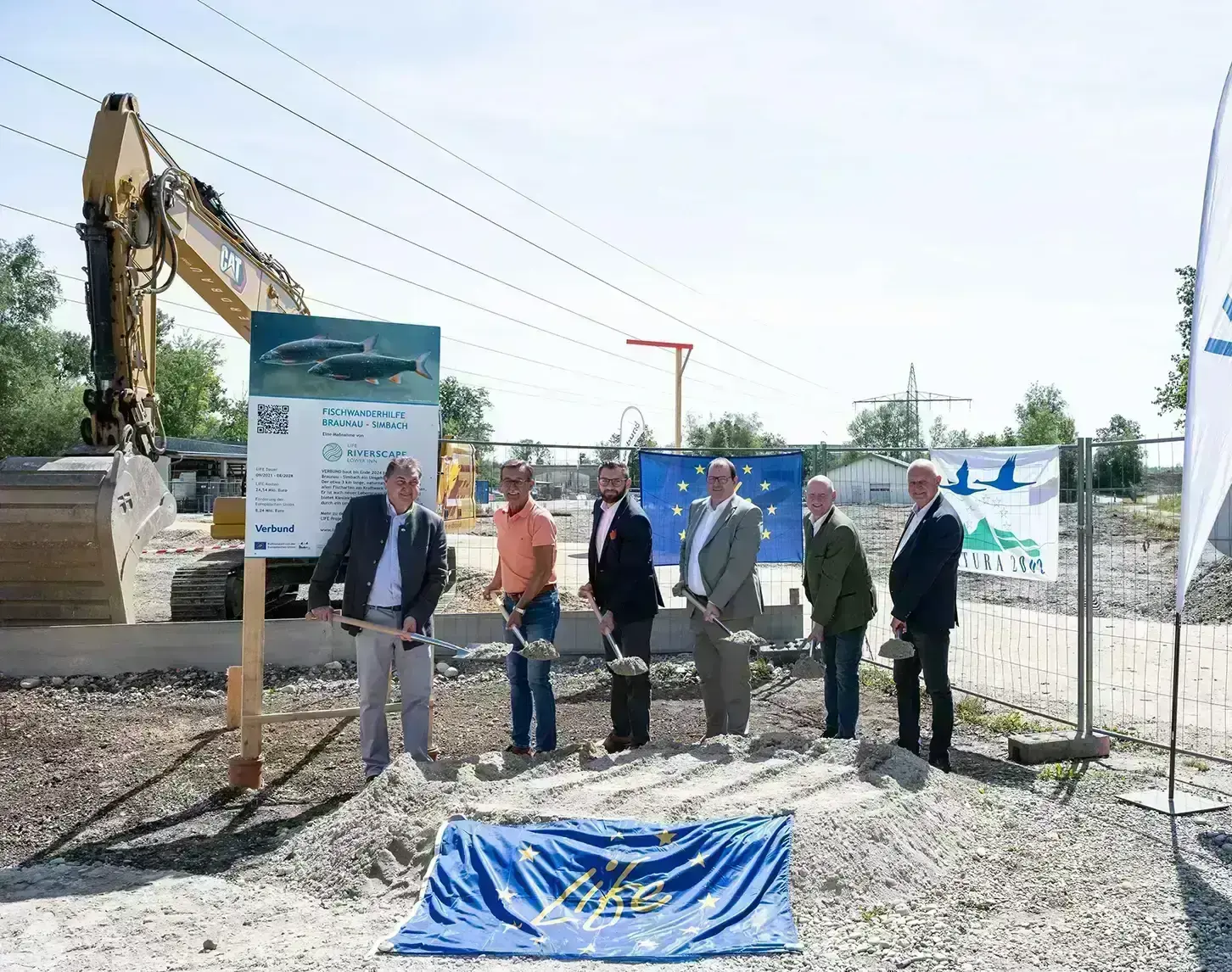 Gruppe von Personen bei einer symbolischen Spatenstichzeremonie an einer Baustelle mit einem Bagger und Informationstafeln im Hintergrund.