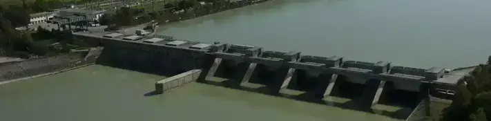 Aerial view of a river power plant with green water, surrounded by a landscape with buildings in the background.