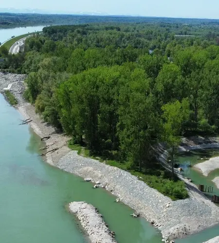 Aerial view of a power plant on a river with backed-up water, adjacent wooded area, and hiking trails in clear weather.