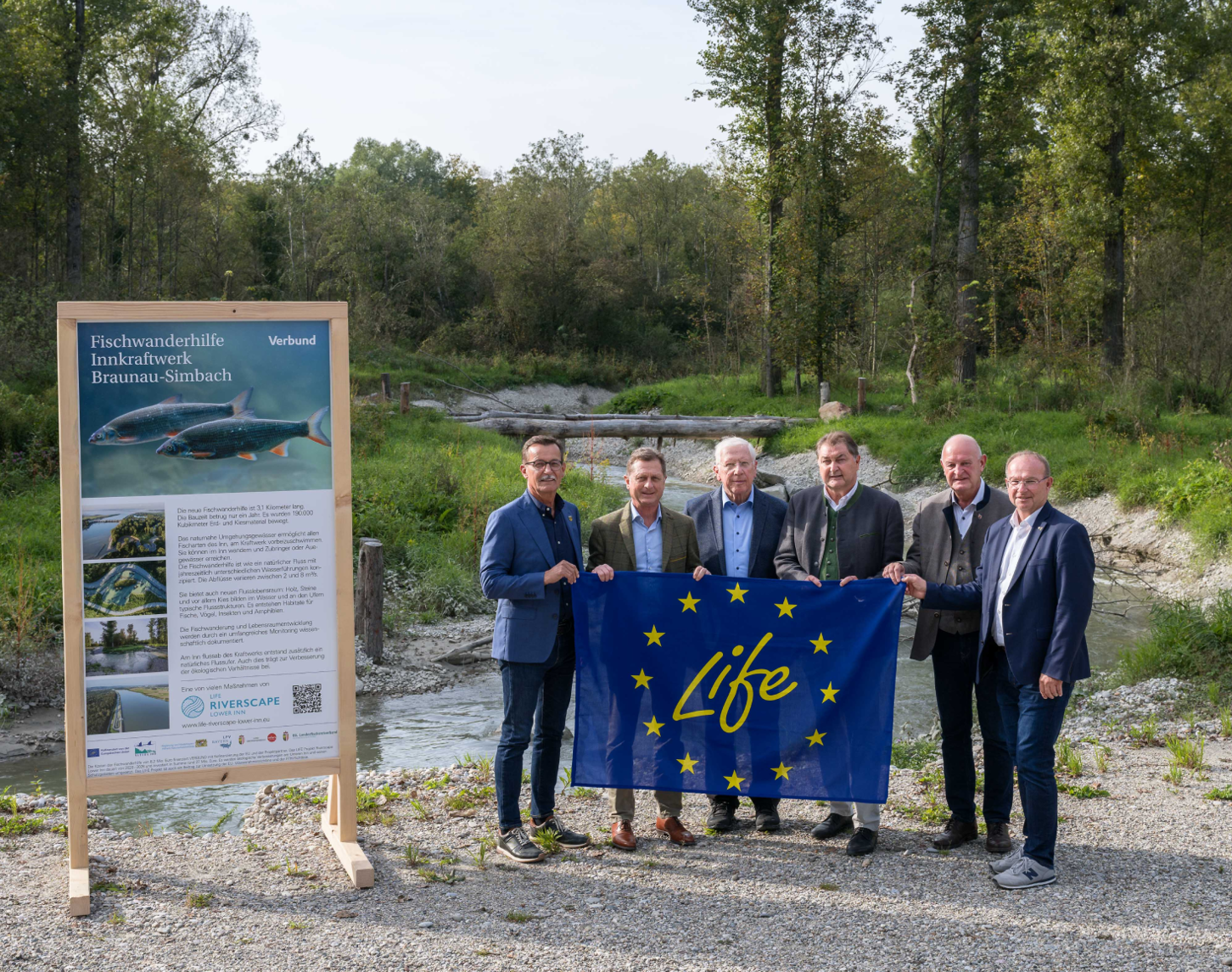 Johann Springer( Bürgermeister von Kirchdorf), Michael Amerer (VERBUND-Wasserkraft Geschäftsführer), Kurt Vallée (stellvertretende Landrat des Landkreises Rottal-Inn), Karl Heinz Gruber (VERBUND-Wasserkraft Geschäftsführer), Klaus Schmid Bürgermeister von Simbach), Johannes Waidbacher (Bürgermeister Braunau) halten gemeinsam die LIFE-Flagge. Im Hintergrund sieht man die Fischwanderhilfe umzingelt von Bäumen in leuchtendem Grün.