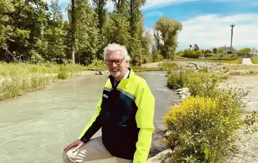 Person in gelber Jacke am Ufer eines fließenden Flusses in einer grünen Landschaft mit Bäumen und Sträuchern im Hintergrund.